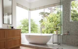 El interior del baño moderno muestra una bañera independiente con ventanas grandes que muestran árboles verdes y plantas en el exterior, con bañera blanca y grifo de agua plateado, ducha gris y una mesa con una planta.