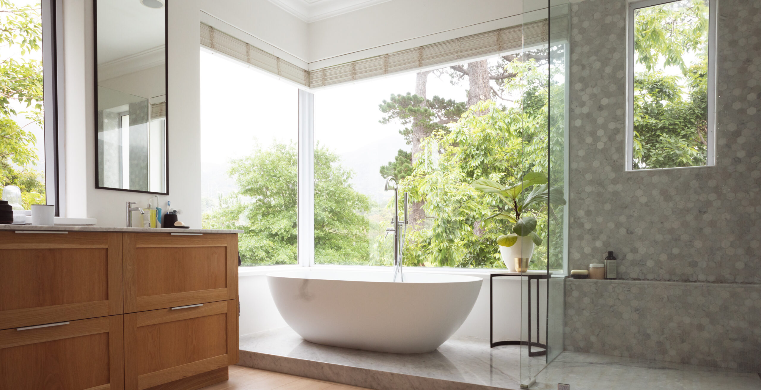 Modern Bathroom Interior Showing Free Standing Bath With Large Windows Showing Green Trees And Plants Outside, With White Bathtub And Silver Water Tap, Grey Shower And A Table With A Plant.